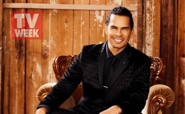 A man in a black patterned suit sits on a vintage chair, smiling, with a wooden backdrop and "TV WEEK" logo in the corner.