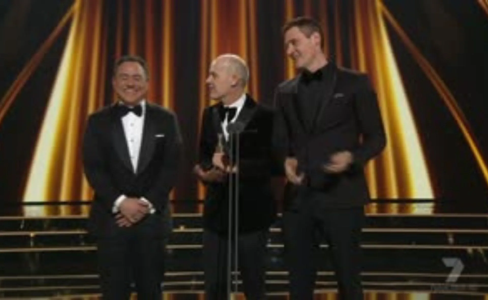Three men in tuxedos stand onstage against a glowing backdrop at an awards event, smiling and holding a trophy.
