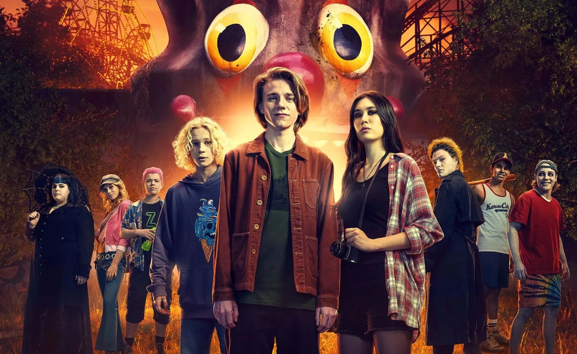 A group of diverse young people stands in front of a giant clown face at an abandoned amusement park, under an orange sky.