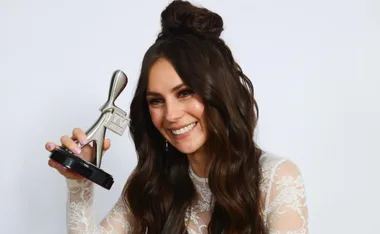 A woman with long hair smiling, holding a silver TV Week award, wearing a white lace top against a plain background.