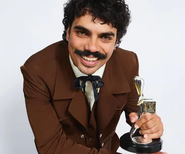Man in a brown suit holding a "TV WEEK" award, smiling at the camera.