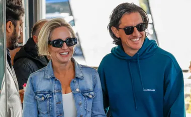 A woman in denim jacket and man in teal hoodie smile, both wearing sunglasses, surrounded by people outdoors.