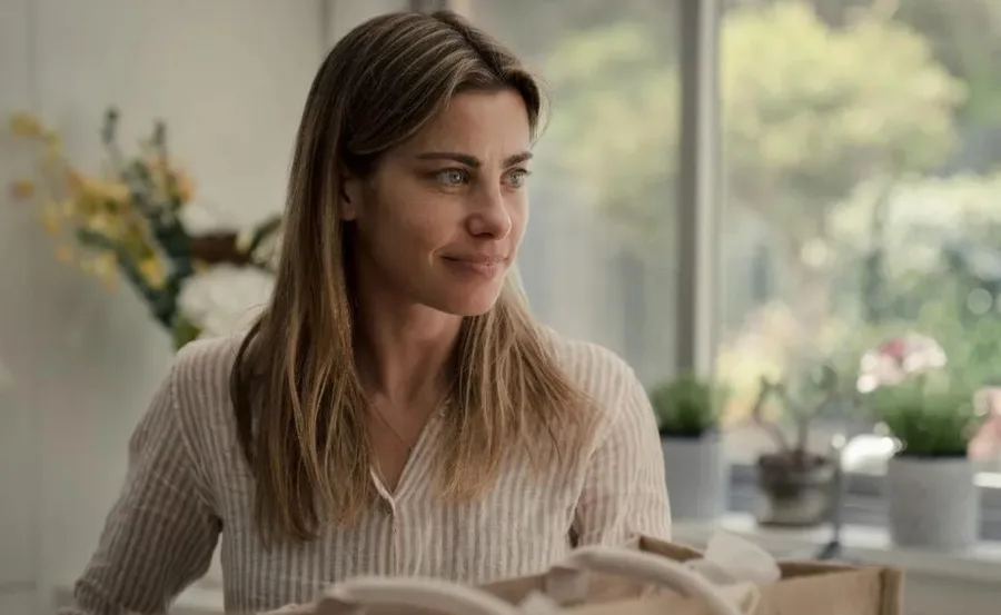 A woman with long hair and a striped shirt smiles indoors, with plants in the background.