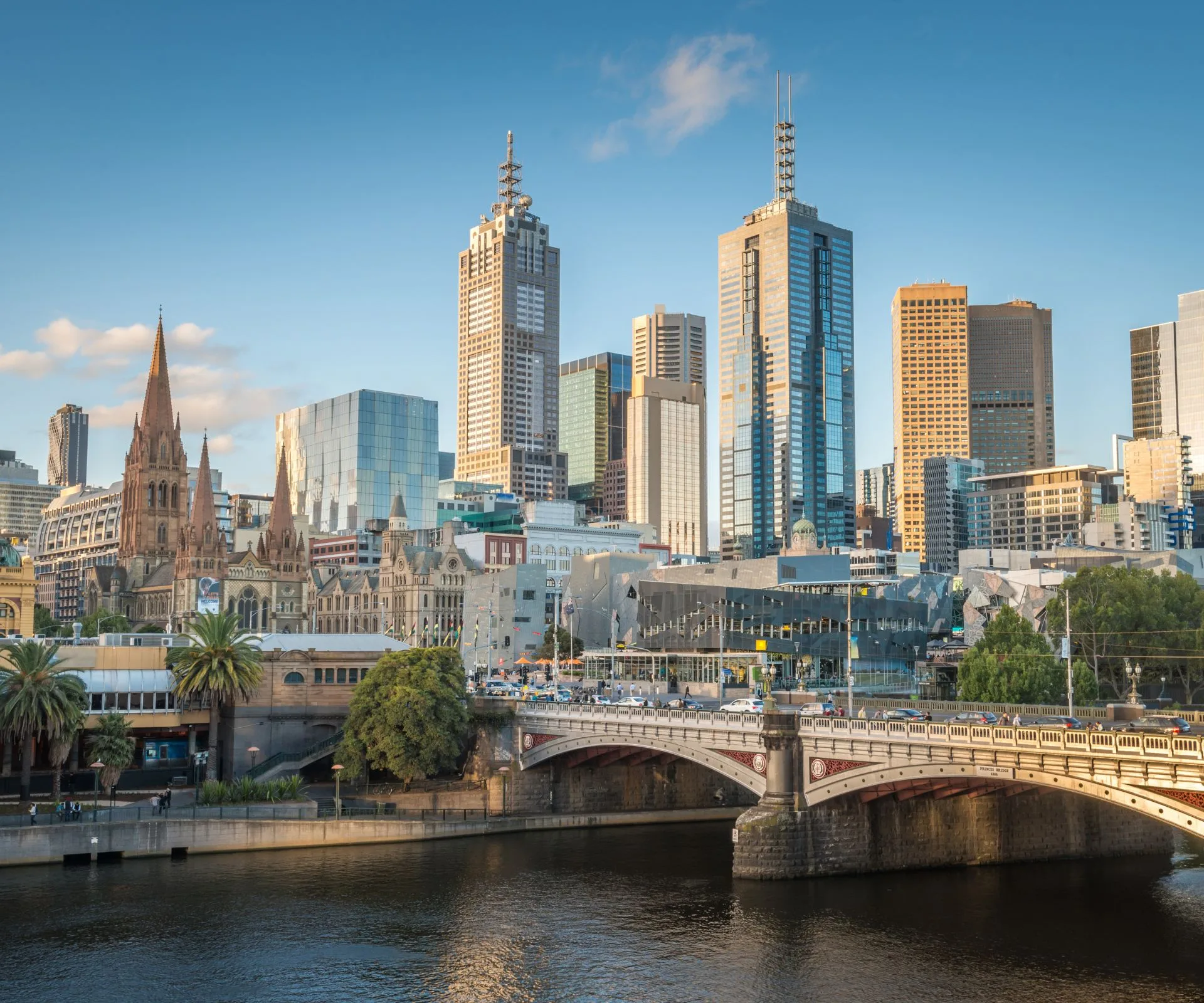 View of Melbourne skyline featuring historic and modern architecture beside a river under a clear blue sky.