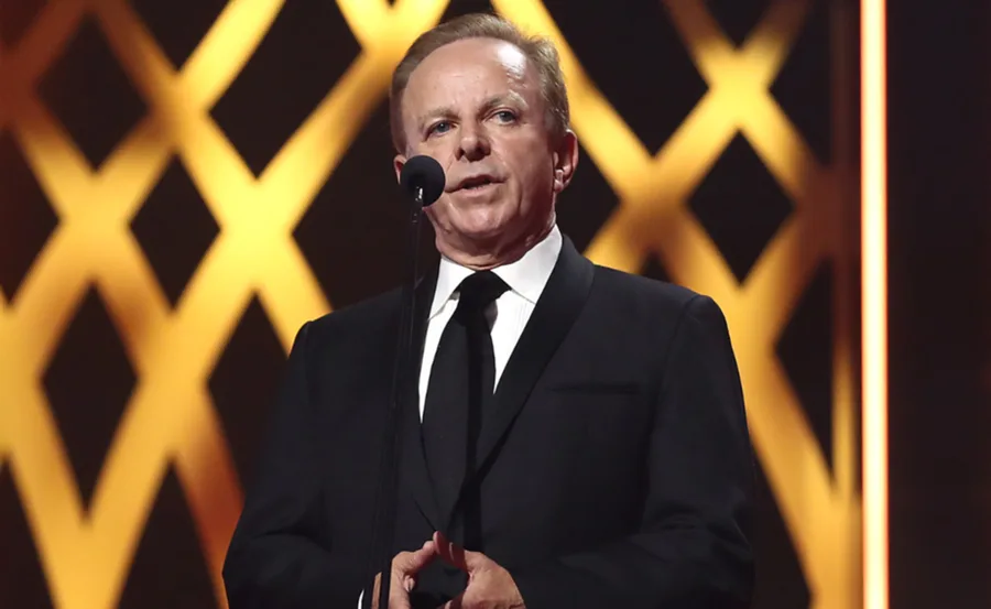 Man in a black suit speaking at a podium, with a patterned golden background.
