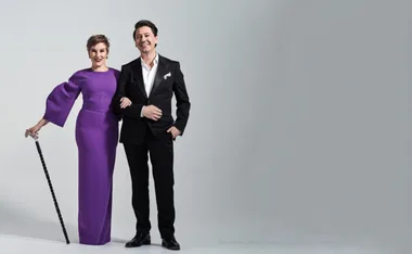 Two people pose together, one in a purple dress with a cane, the other in a black suit.