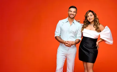 Two people smiling, one in a light blue outfit and the other in a white and black dress, against an orange background.