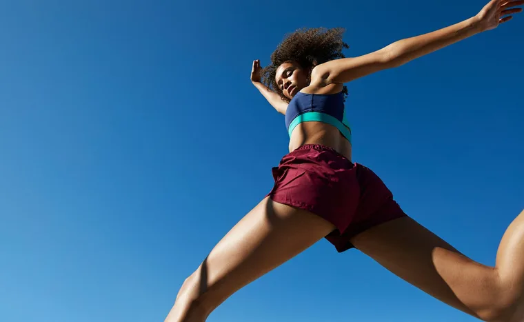 A woman in athletic wear jumps against a clear blue sky, arms reaching upward.
