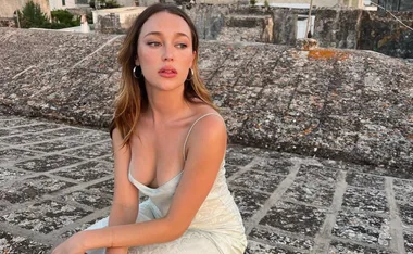 Young woman in a light dress sitting on a stone rooftop, gazing thoughtfully to the side.