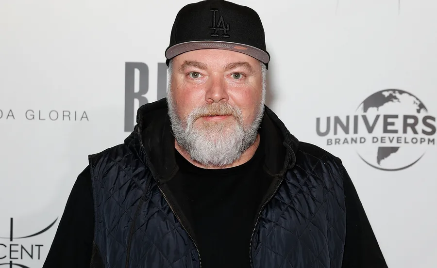 A man with a beard wearing a black cap and quilted jacket stands in front of a branded event backdrop.