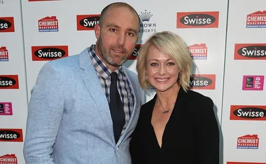 Man in a light blue suit and woman in black dress posing at a Swisse event backdrop.