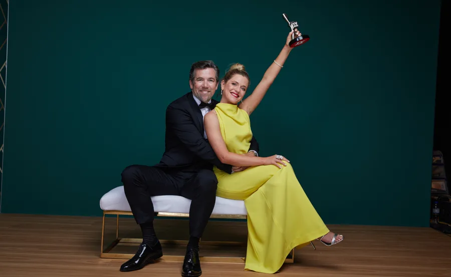 Couple on a bench, woman in yellow dress holds up award, with man in suit hugging her, smiling against a teal background.