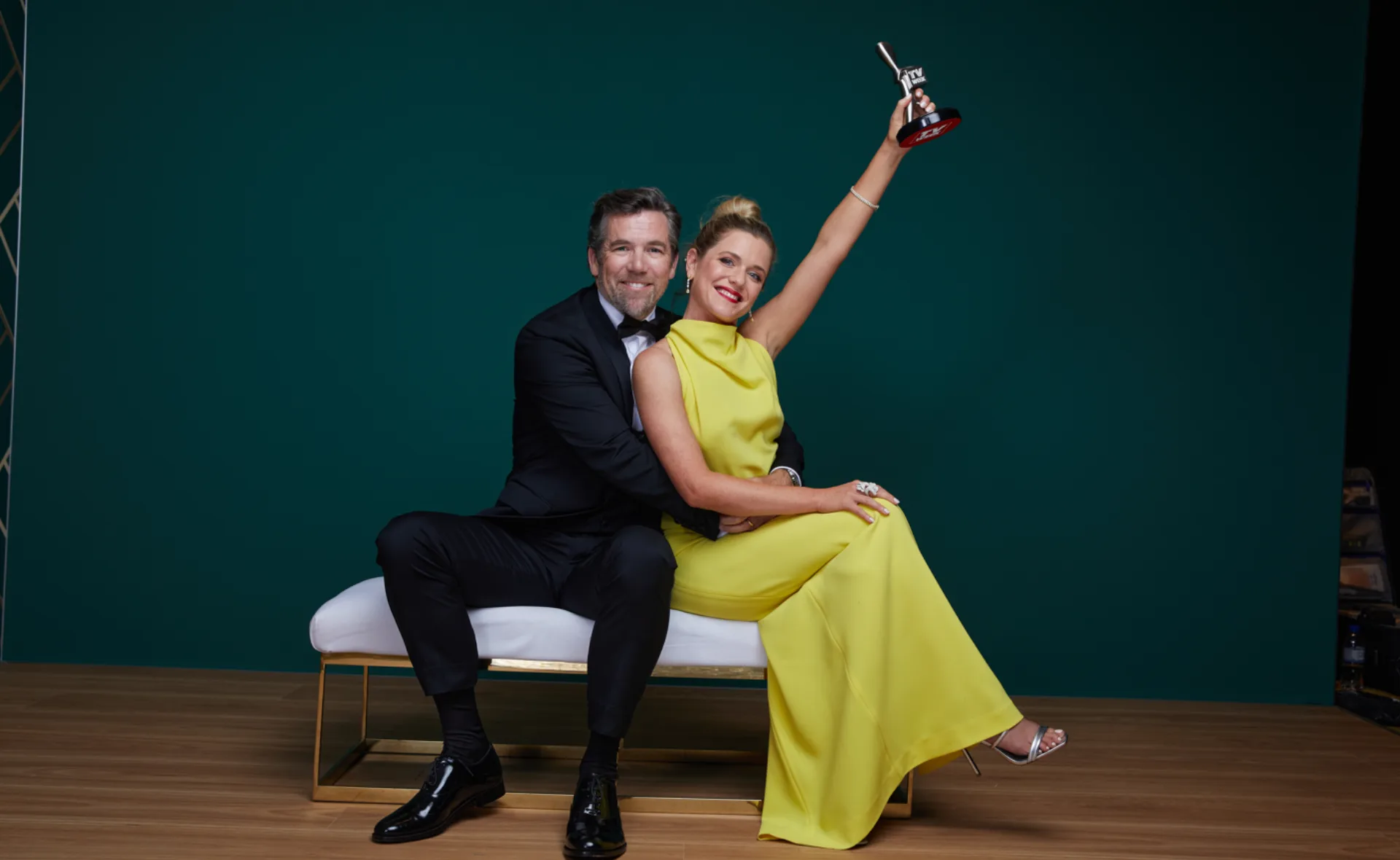 Couple on a bench, woman in yellow dress holds up award, with man in suit hugging her, smiling against a teal background.