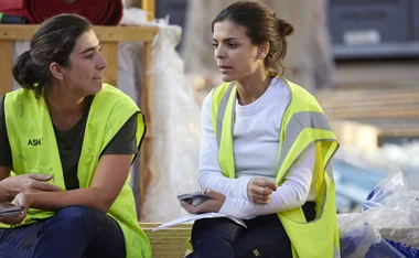 Two women in yellow safety vests sit and talk at a construction site, holding phones and papers.