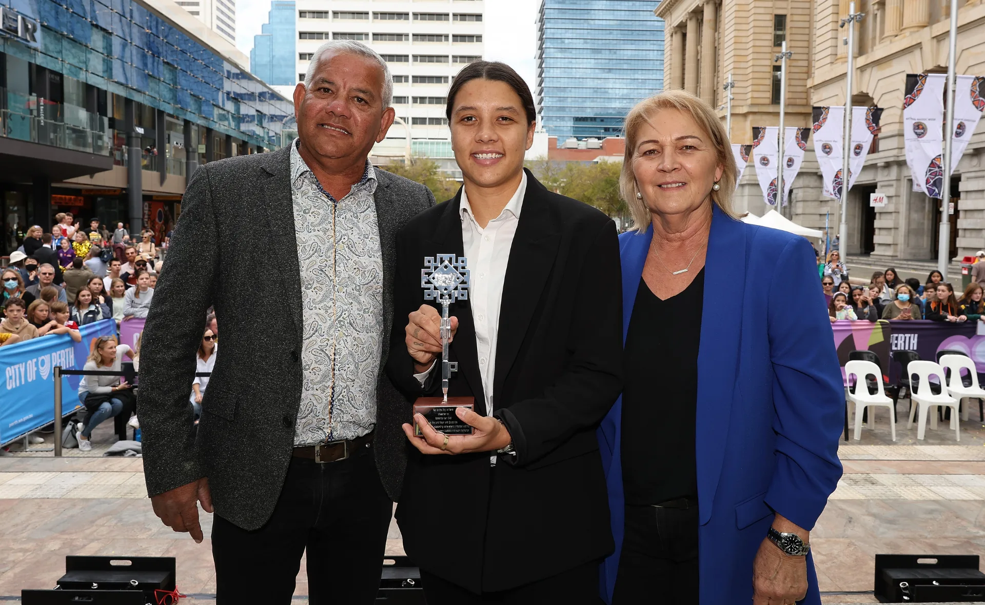 Three people posing outdoors in front of a crowd; the center person holds a trophy.