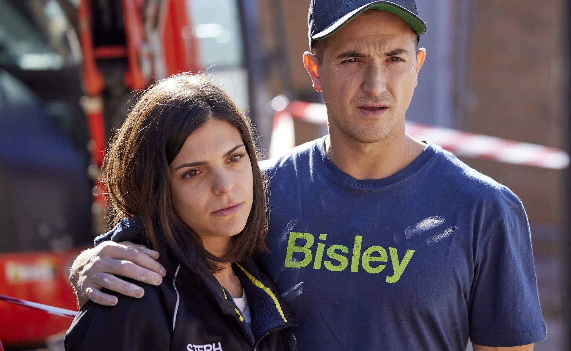 A woman with "Steph" on her jacket, leaning on a man in a "Bisley" shirt, standing outdoors near construction equipment.