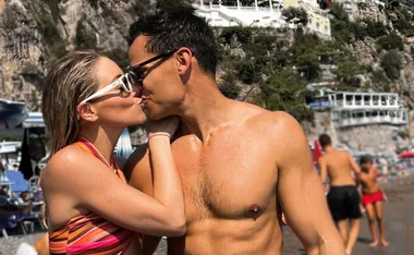 Couple kissing on a sunny beach, with cliffs and buildings in the background.