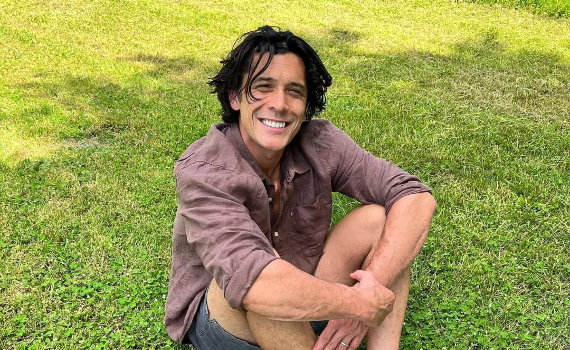 A person with dark hair sits on grass, wearing a maroon shirt and shorts, smiling broadly at the camera.