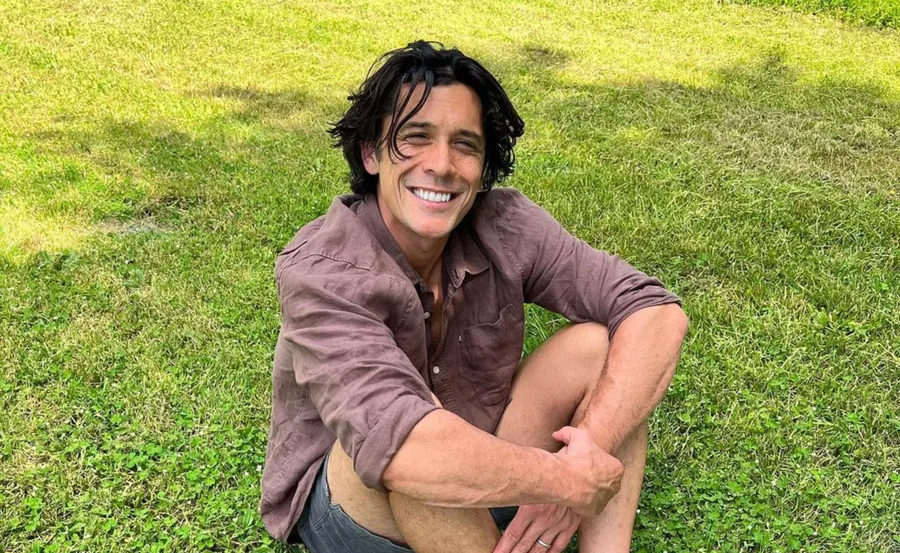 A person with dark hair sits on grass, wearing a maroon shirt and shorts, smiling broadly at the camera.