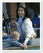 A woman with long dark hair holding a baby by the edge of a pool, both wearing light clothing, on a sunny day.