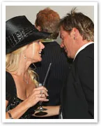 A woman in a black hat and dress is smiling and talking to a man in a suit at an event.