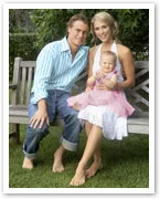 A family sits on a garden bench; a man and woman with a toddler in a pink dress on the woman's lap, all smiling.