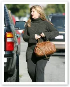 A woman walking outdoors, holding a large brown bag, wearing a dark sweater and pants, with parked cars in the background.