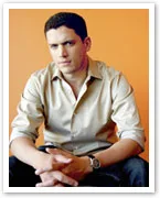 Wentworth Miller in a light shirt sits against an orange background, hands clasped, wearing a watch.