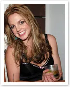 A woman with long hair smiling and holding a drink, wearing a black outfit, against a neutral background.