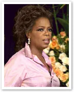 Oprah Winfrey in a pink blouse speaks on stage, with a background of flowers.