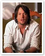 A person with shoulder-length hair wearing a striped shirt sits in a wooden chair, looking ahead.