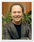 Billy Crystal smiling, wearing a dark jacket, standing in front of green plants.
