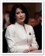 A woman in a white jacket with dark hair, looking thoughtful with a crowd in the background.