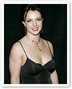 A woman in a black dress smiles, wearing a necklace with red accents, against a dark background.