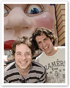 Two smiling men stand in front of a large statue of a face with exaggerated features in a playful setting.