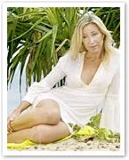 A woman in a white dress sitting on sand near tropical plants, looking thoughtful.