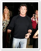 A man in a black shirt and jeans stands outdoors, with people around, one with a camera and others looking towards him.