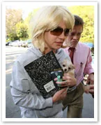 Person in sunglasses holding a small dog and a notebook, accompanied by another person outdoors.