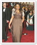 A person in a brown dress with silver sleeves walks the red carpet at a formal event, with a crowd in the background.