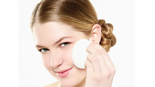 Woman using a round makeup sponge on her face, smiling, with hair styled in a bun.