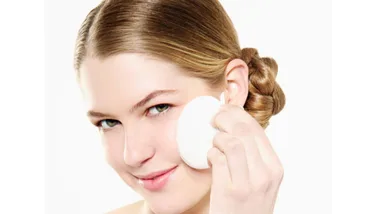 Woman using a round makeup sponge on her face, smiling, with hair styled in a bun.