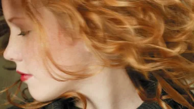 Red-haired person with loose curls and closed eyes, head turned to the side, wearing a dark top.