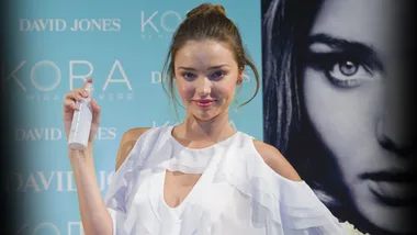 Miranda Kerr holding a product from her KORA Organics range at a launch event, smiling in front of branded display.