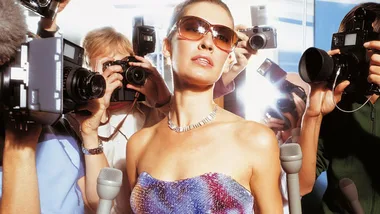 Celebrity posing confidently in sunglasses as paparazzi take photos on the red carpet.