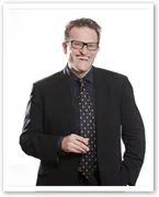 A man in a suit with glasses, smiling and holding an object, stands against a white background.