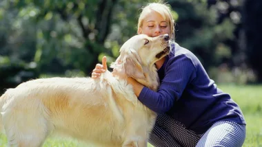 A woman lovingly hugs a golden retriever in a sunny park.