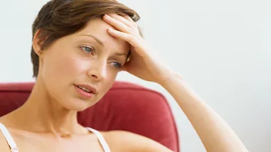 A woman sitting on a red chair, her hand on her forehead, looking pensive or thoughtful.