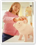 A child putting a coin into a pink piggy bank on a table, teaching the importance of saving money.