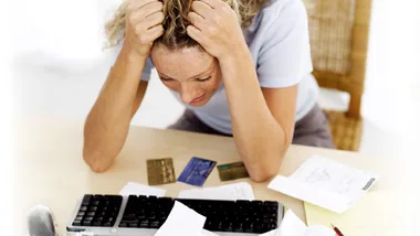 A stressed woman holding her head at a desk cluttered with bills, credit cards, and a keyboard.