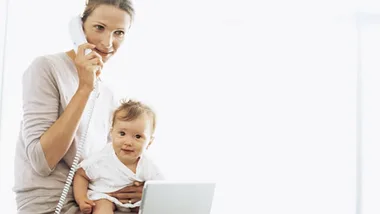 Mom holding baby while talking on corded phone and using a laptop, both looking at camera.